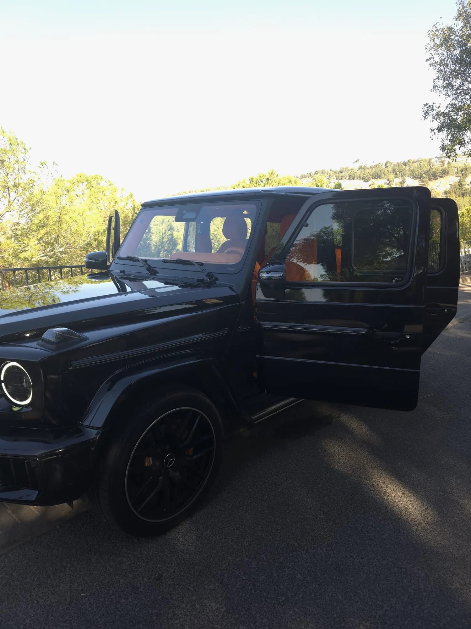 Black Mercedes-AMG G63 from the front three-quarter, driver's door open to orange leather seats, wooded hills behind